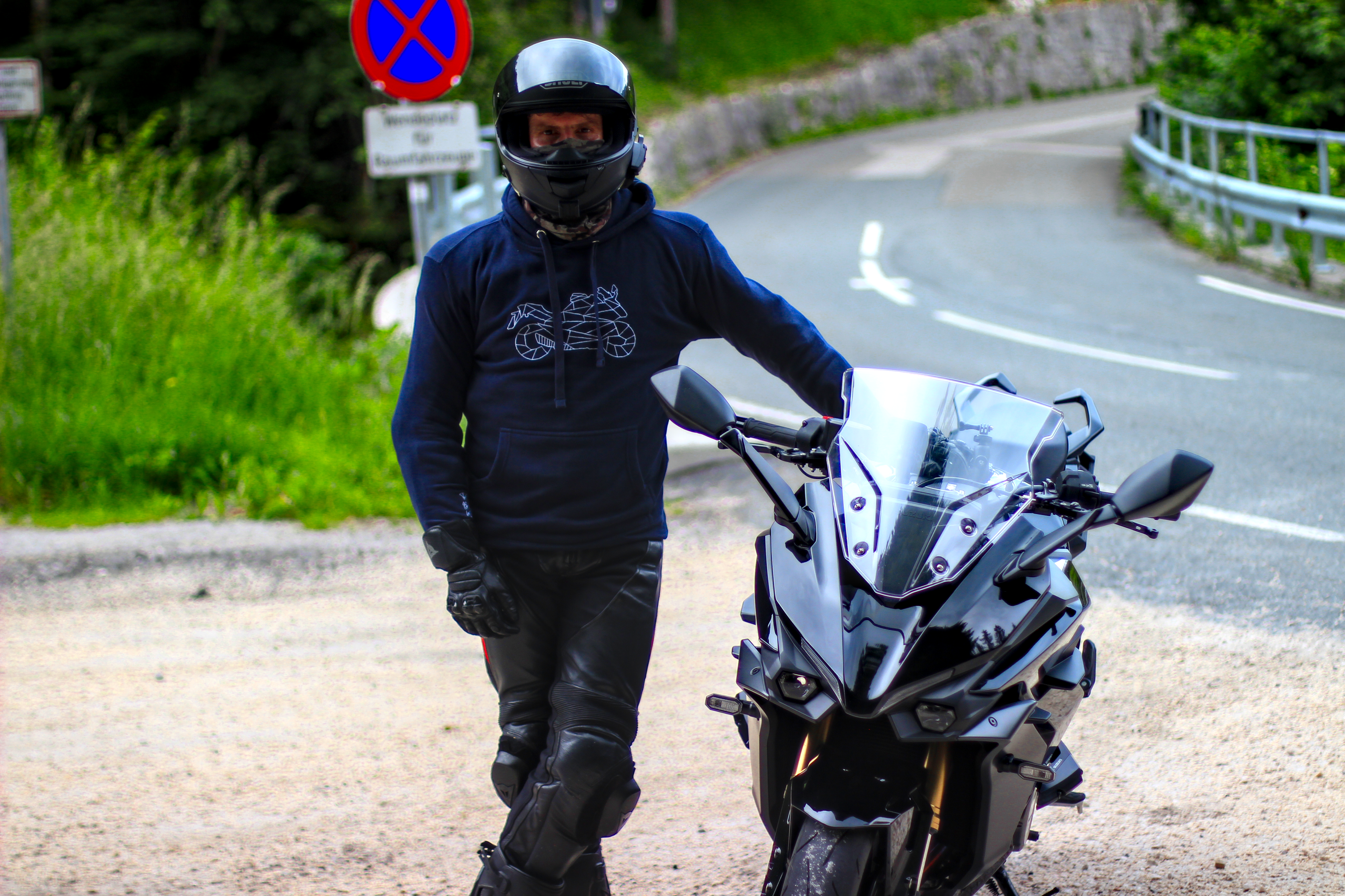 Man wearing a black motorcycle style hoodie standing next to a black sport motorcycle on a winding road