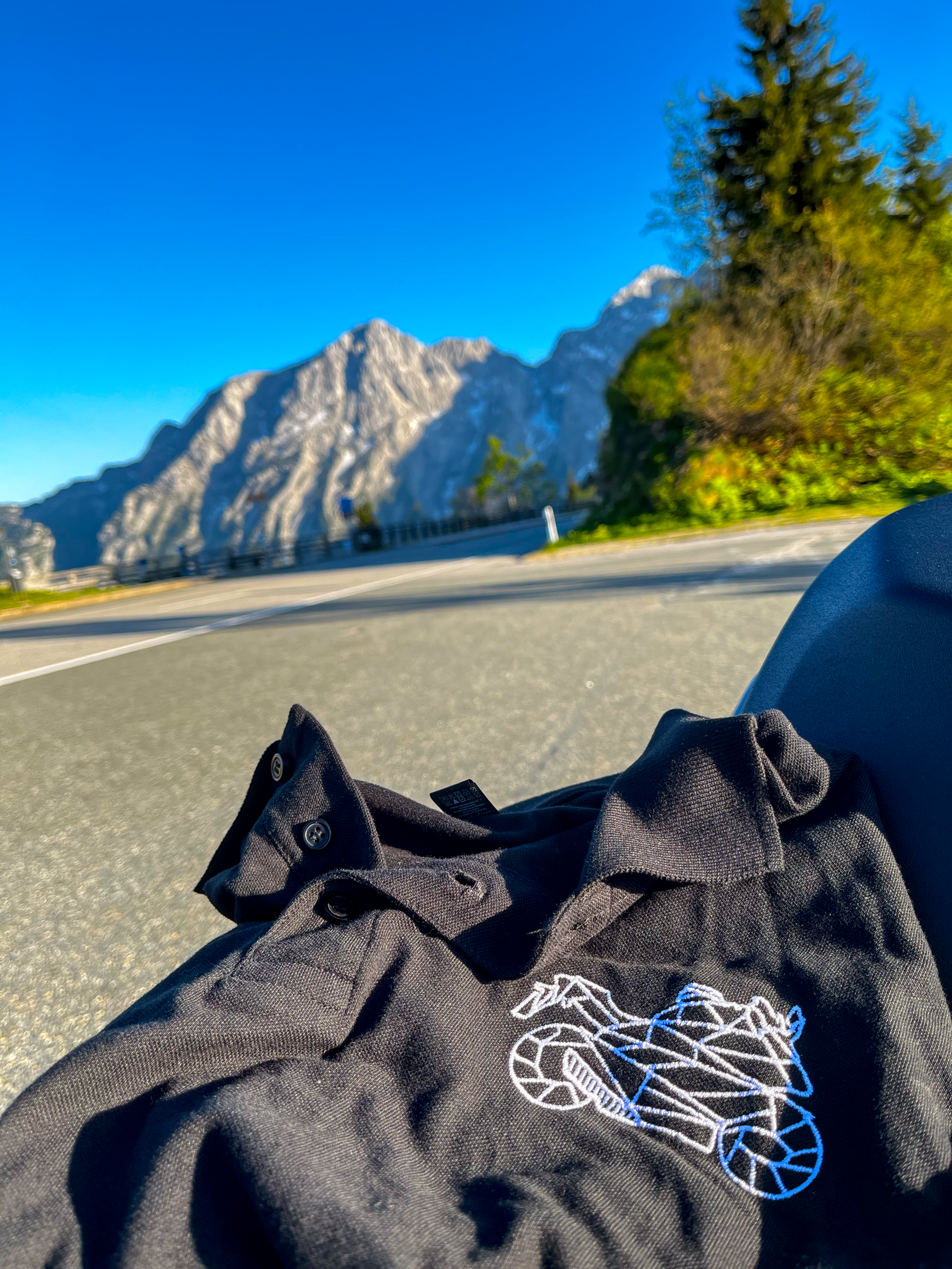 Black motorcycle polo shirt for men with embroidered bike design laid on motorcycle seat by mountain road