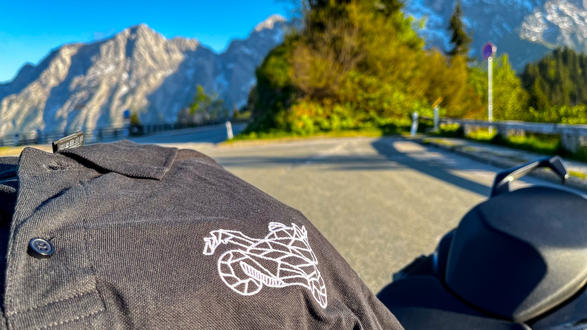 The Biker Mule black motorcycle polo shirt men with white logo displayed on scenic mountain road
