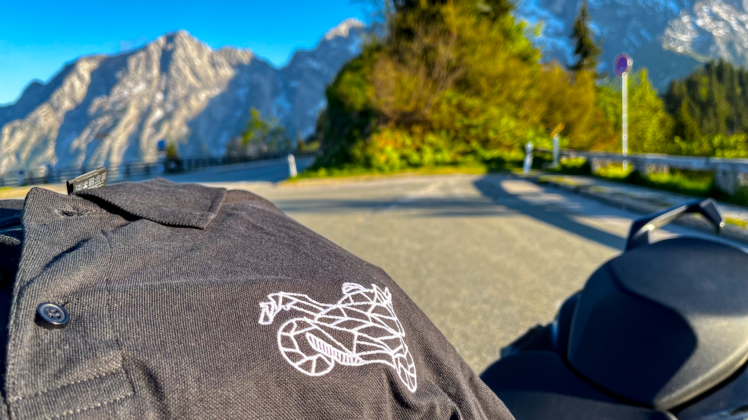 The Biker Mule black motorcycle polo shirt men with white logo displayed on scenic mountain road