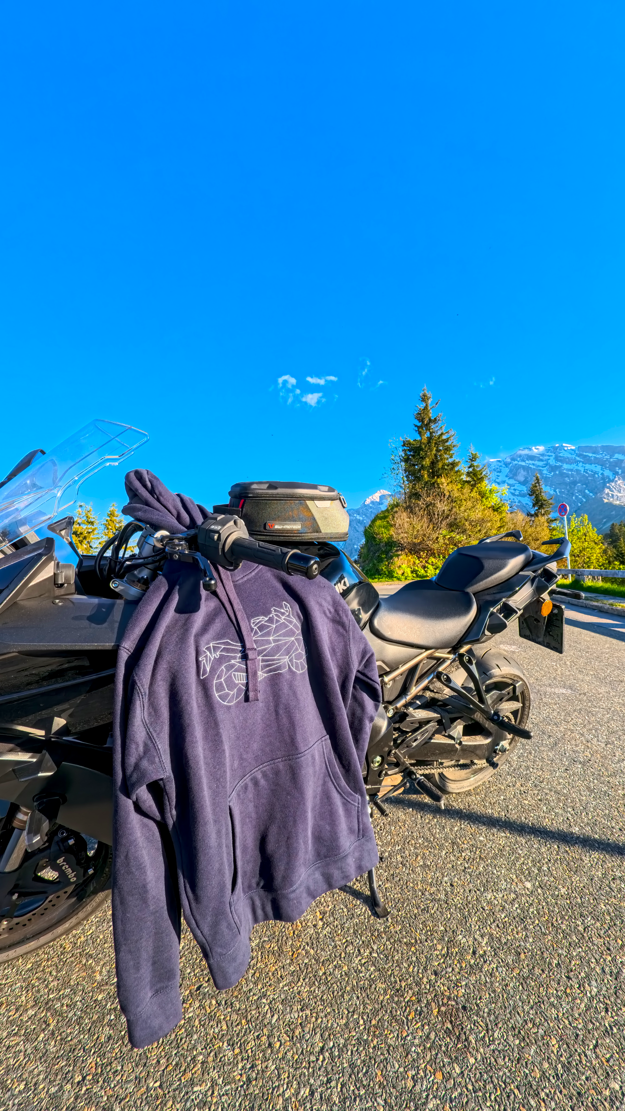 The Biker Mule unisex biker hoodie hanging on a black motorcycle with clear blue sky and mountains in the background